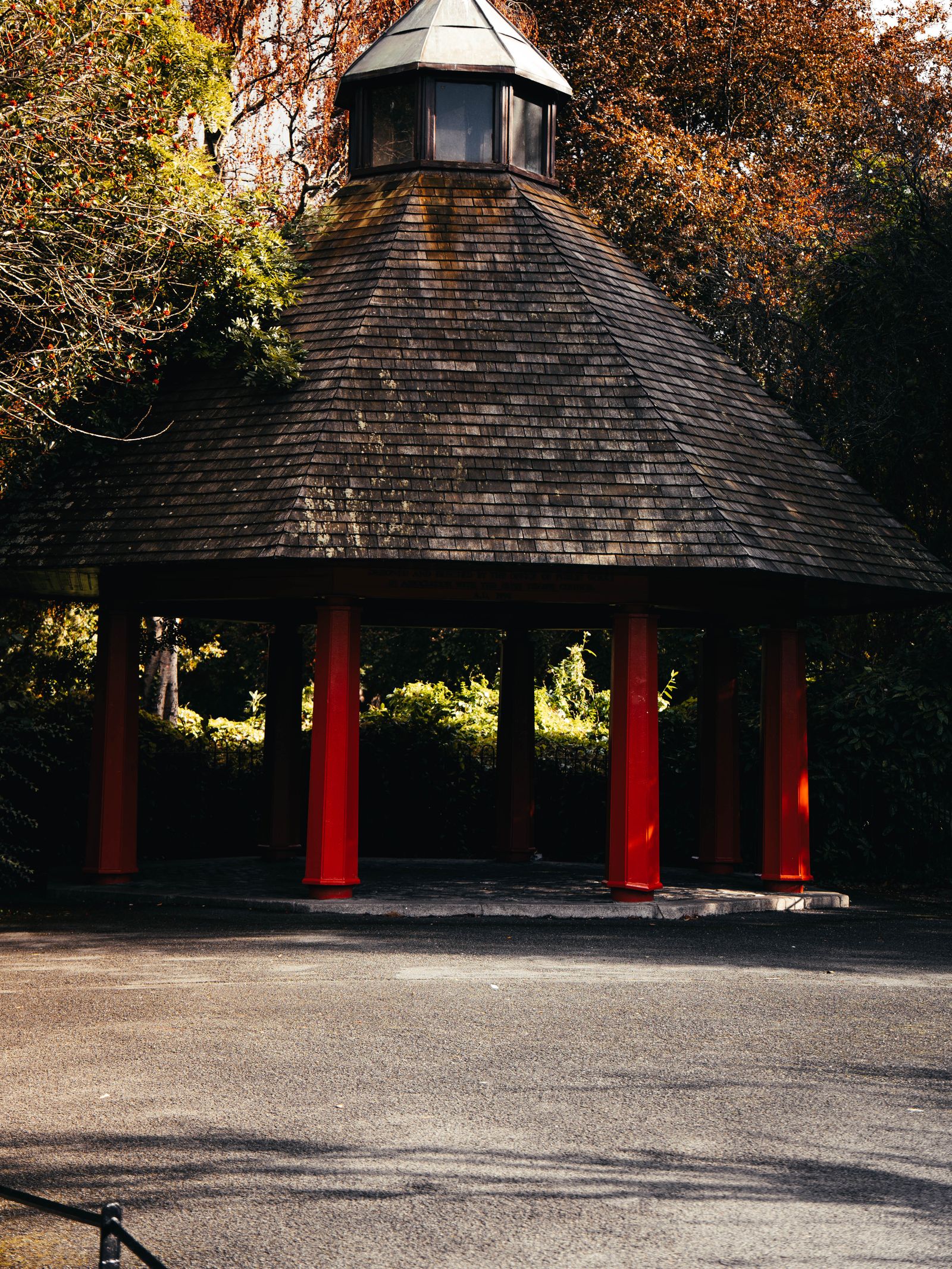 A wooden pagoda-style shelter with red pillars in a Dublin park, autumn trees behind.