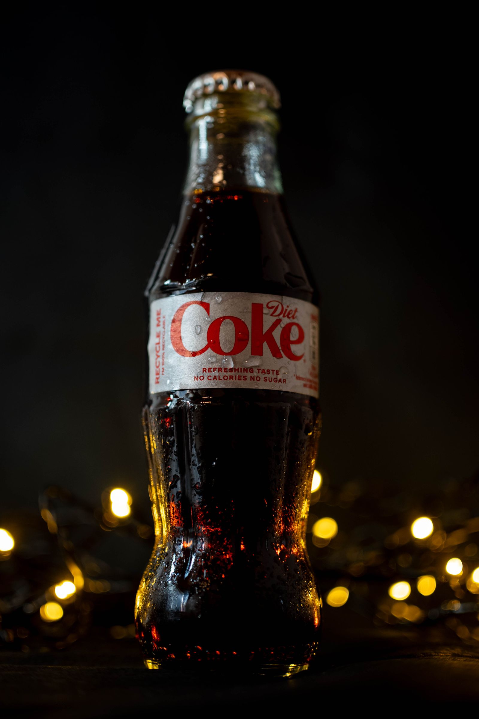 A Diet Coke bottle in front of warm out-of-focus fairy lights.