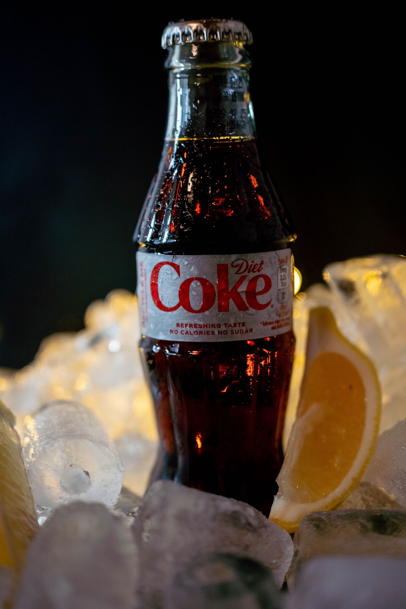 A Diet Coke bottle in crushed ice with a lemon wedge, against black.