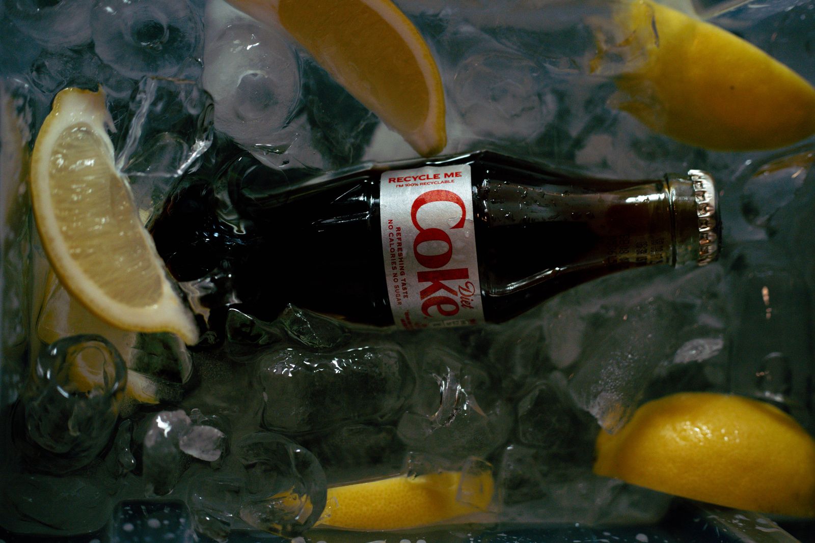 A Diet Coke glass bottle lying in ice with lemon wedges.