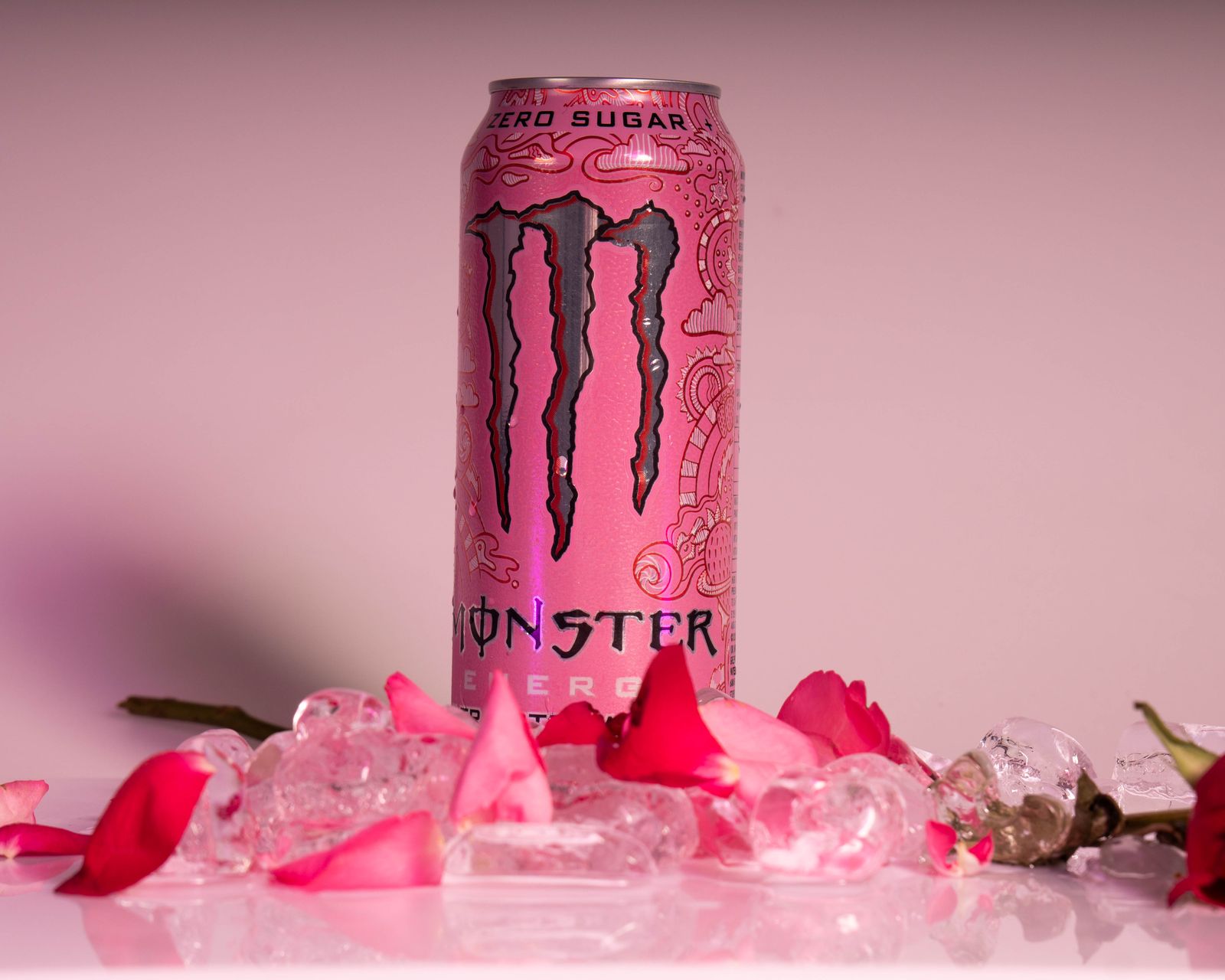 A pink Monster Energy Ultra can standing on ice and rose petals against a pink background.