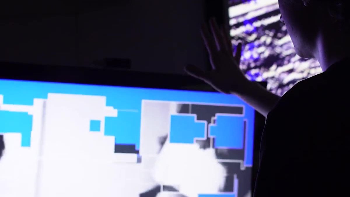 Hands pressed against a screen in a dark room, the image breaking into black-and-white blocks.